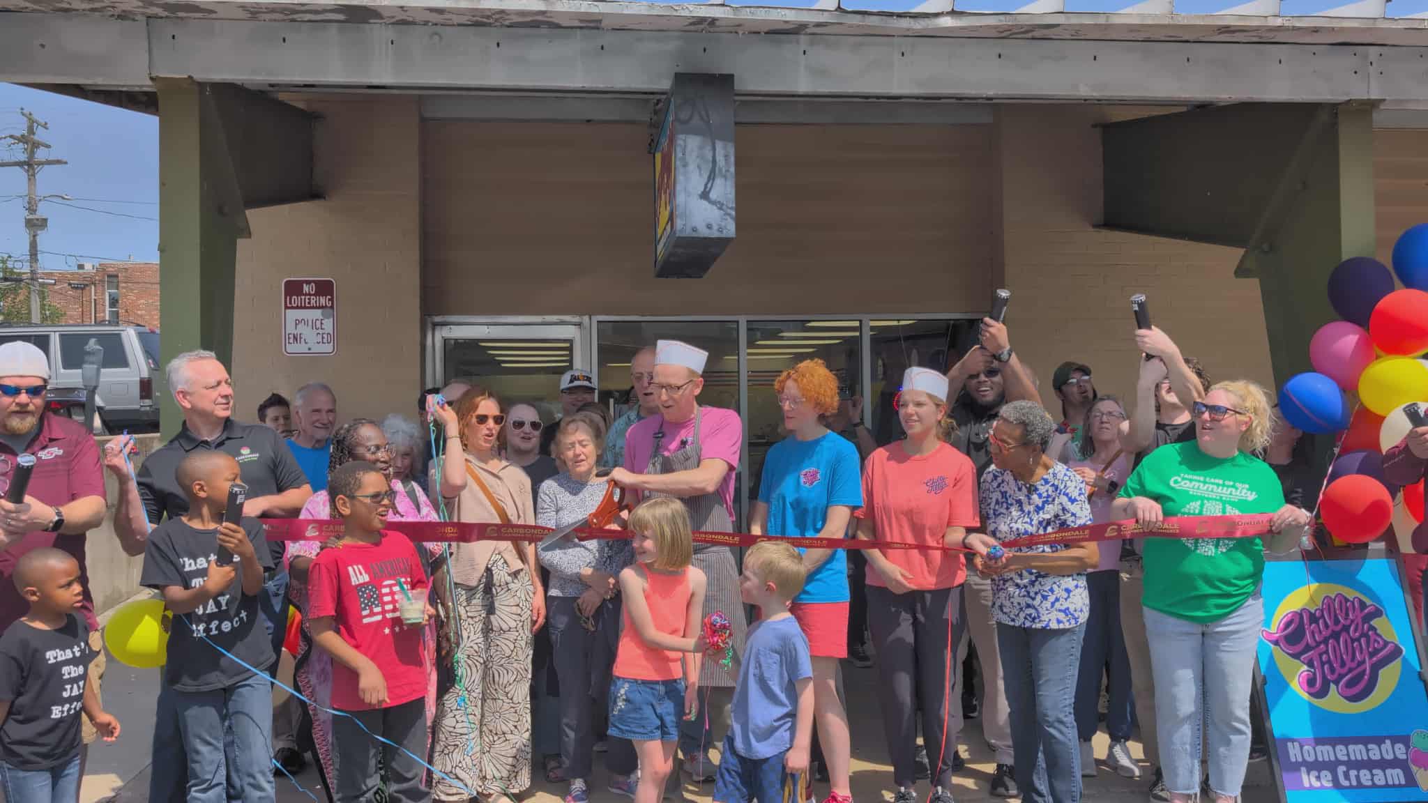 Several people stand in front of Chilly Jilly's main entrance for a ribbon cutting. The owner is just about to cut the ribbon and everyone in the crowd is watching with anticipation. Several people in the audience have confetti launchers. 