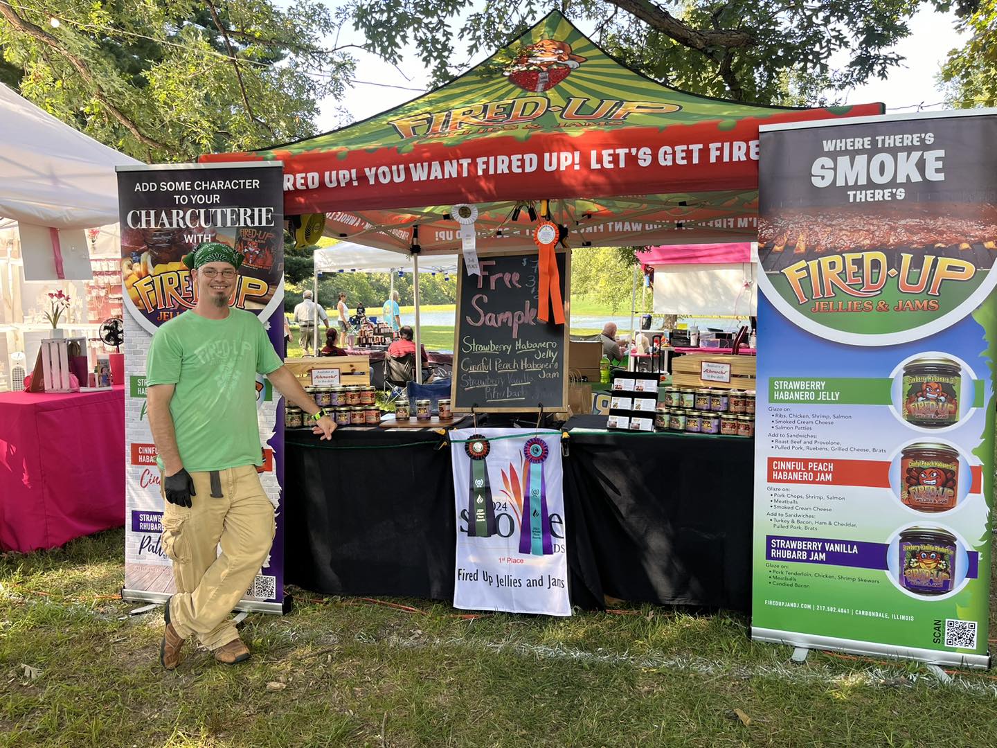 A booth at a vendor fair. The pop-up tent is branded "Fired Up Jellies and Jams." The table below is has jelly and jam displays on it. There's a chalkboard in the center that says "free samples." There is a large, vinyl display banner on either side of the table with Fired Up branding. Owner Michael Smith is standing off to the left in a green Fired Up t-shirt. 