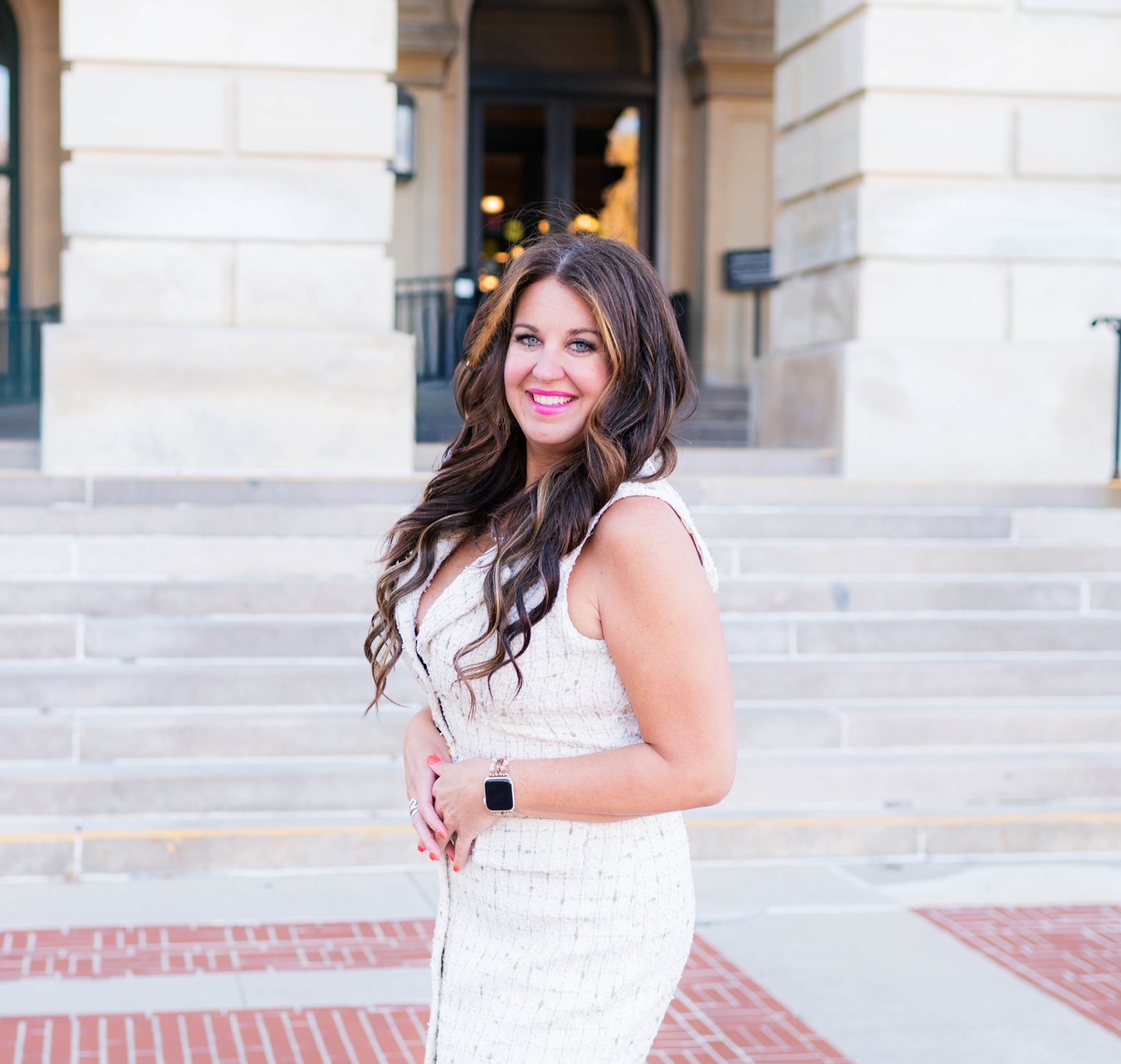 Sarah Garcia, a white woman with long brown hair and blue eyes, stands in front of a building with large, white, square columns and white stairs. Sarah is wearing a white dress that is of similar color. The red brick of the walkway provides contrast in the picture.  