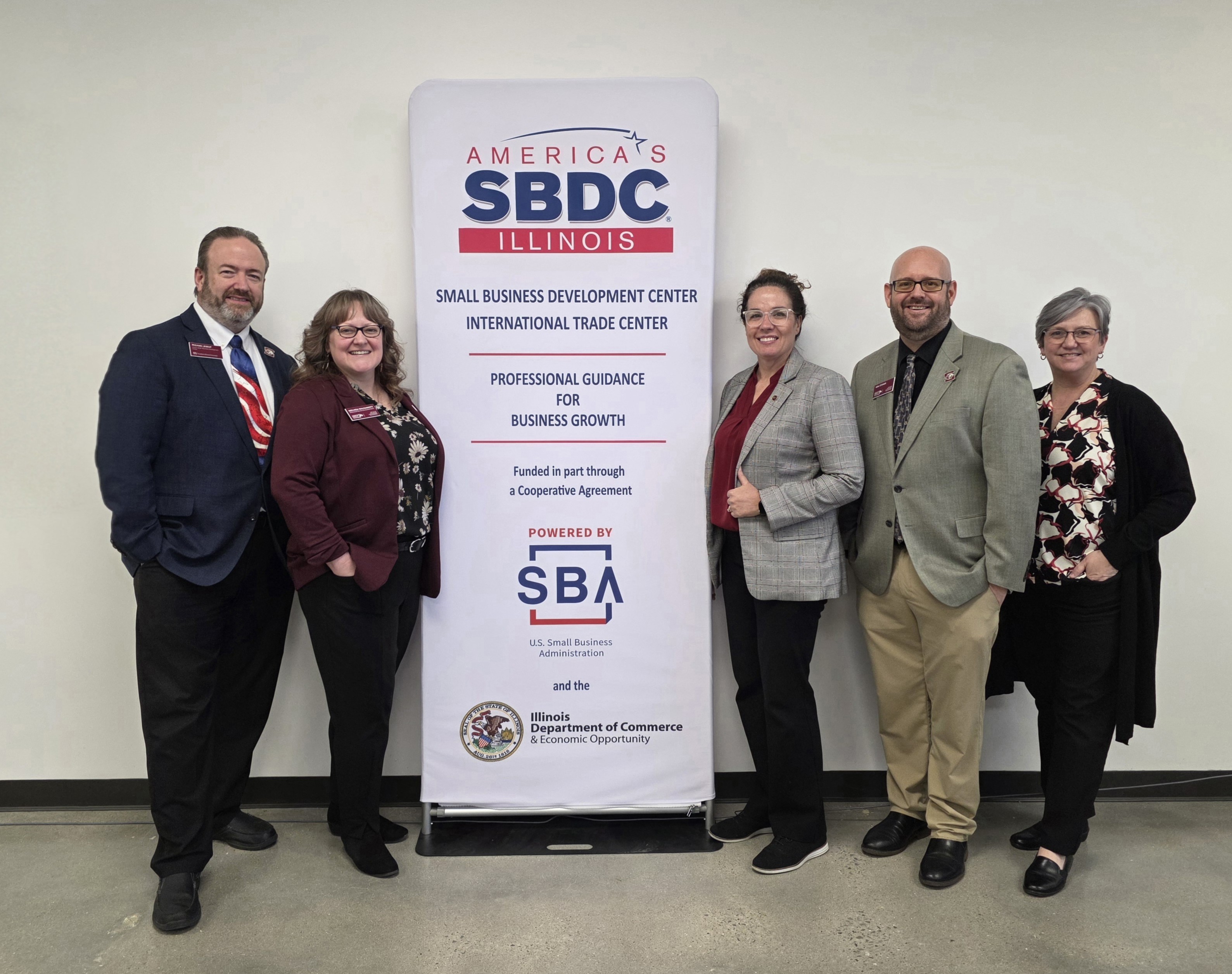 The Illinois SBDC at SIU staff stands on either side of a 7' banner stand with the SBDC logo on it. They are dressed in business attire and smiling. 