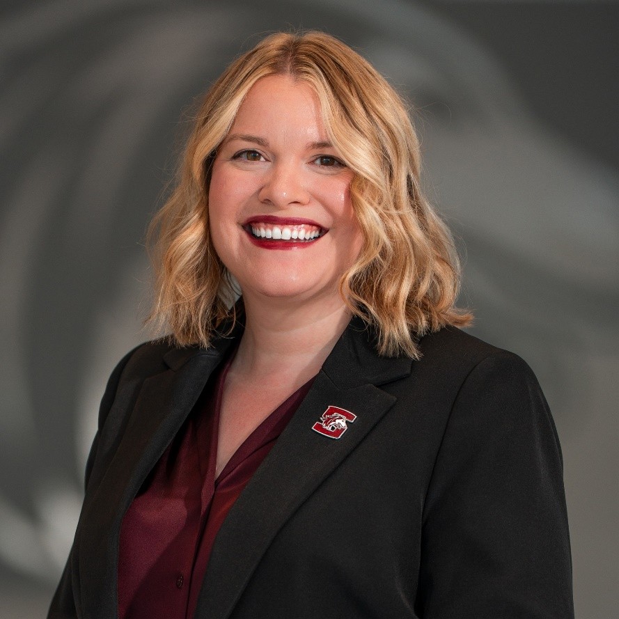 A headshot of a white woman with shoulder-length, wavy, blond hair. She is wearing a maroon shirt under a black suit jacket with a SIU lapel pin.