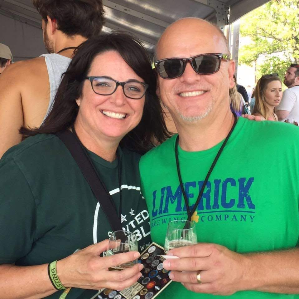 A couple stands side by side with an arm around the other. They are dressed in green shirts and holding beverages. There is a crowd in the background. 