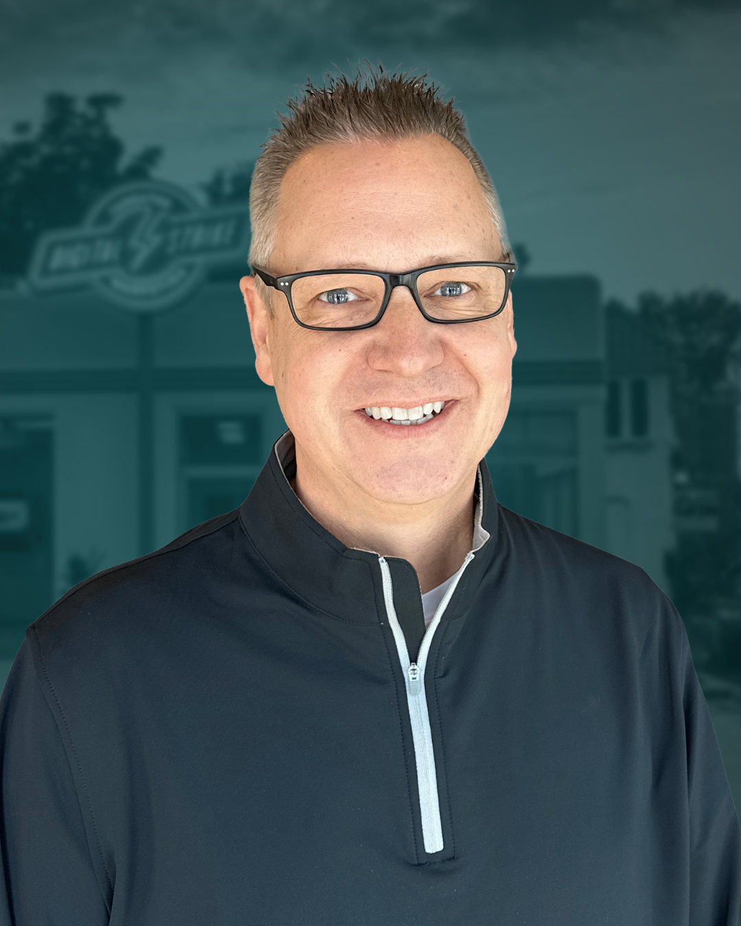 A headshot of a white man with gray hair, blue eyes, and black framed glasses. He is wearing a charcoal-colored three-quarter zip pull over. The background is a blurred picture of the Digital Strike building with a blue filter over it.