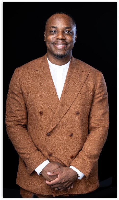 A black man stands with his hands clasped in front of him. He is wearing a white shirt under a brown, double-breasted suit jacket. 