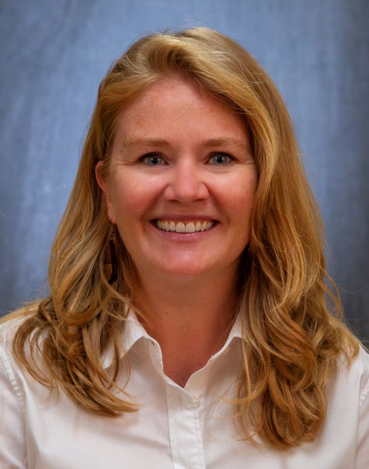 A headshot of an white woman with long, blond hair. She is wearing a white, button up shirt. There is a blue background behind her. 