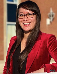 A headshot of an Asian woman with long, dark hair. She is wearing black-framed glasses and a black shirt under a red suit jacket.
