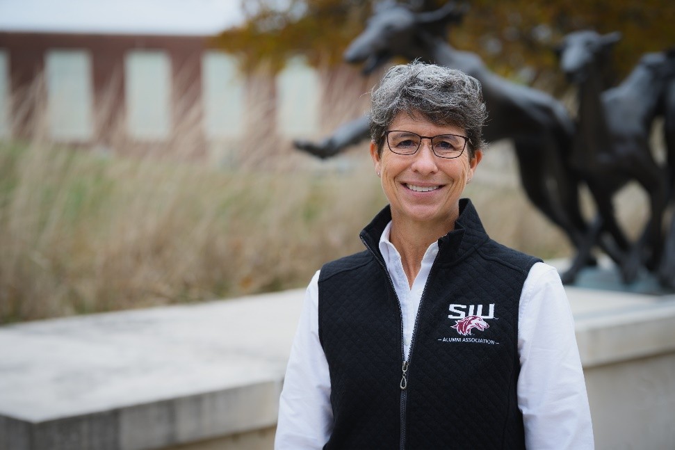 A white woman with short gray hair and glasses stands in front of a Saluki statue. She is wearing a white dress shirt under a black, SIU-branded zip-up vest. 