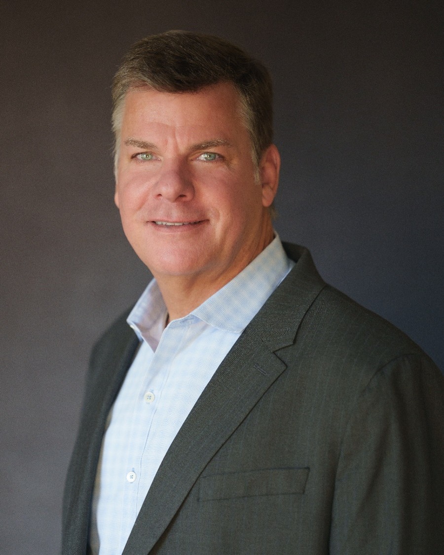 A headshot of a white man with graying hair in a blue and white gingham shirt under a gray suit jacket.