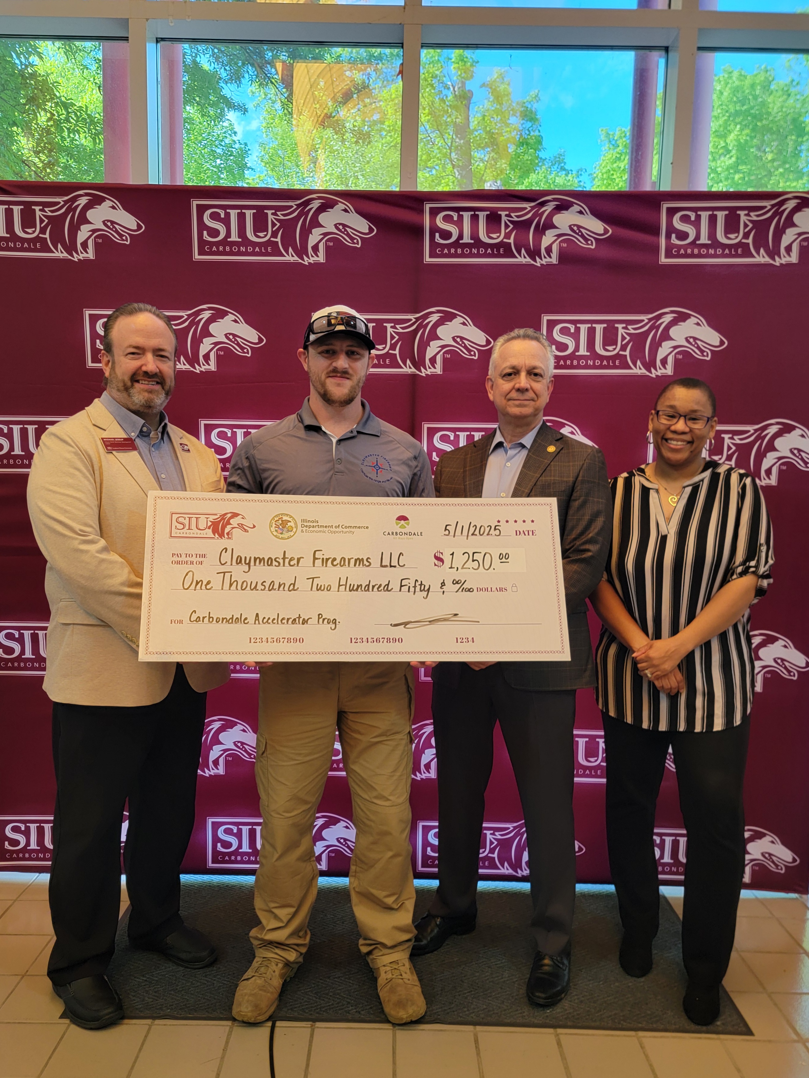 Matthew Huff, owner of Claymaster Firearms, holds a ceremonial check at the Carbondale Accelerator Program graduation ceremony. SBDC instructors and Steve Mitchell from the City of Carbondale stand on either side of him. 