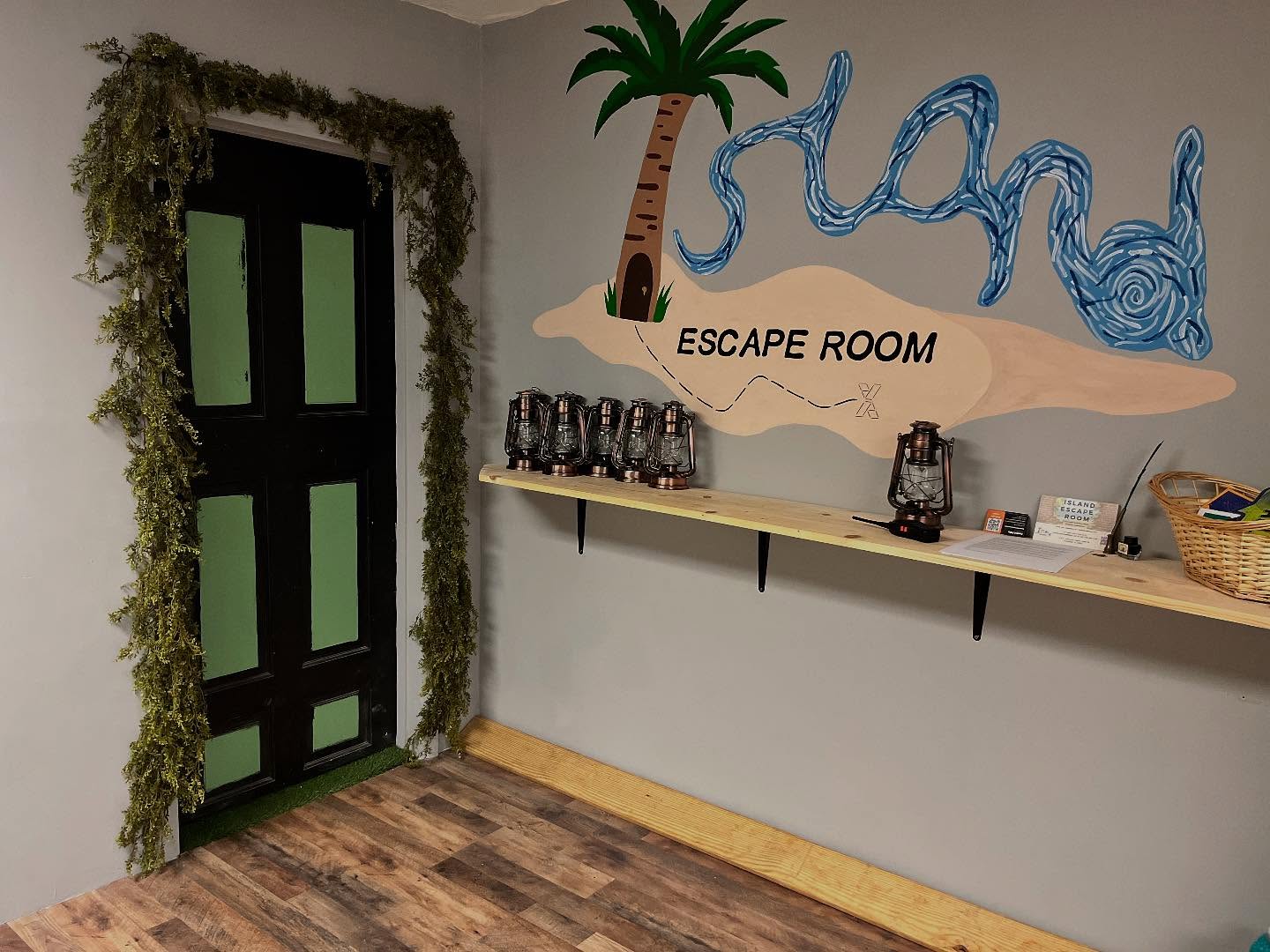 I picture of the door that leads to the first puzzle room in Island Escape Room. There is a shelf with camping lanterns and there is fake foilage stuck to the wall to suggest you are in the forrest. 