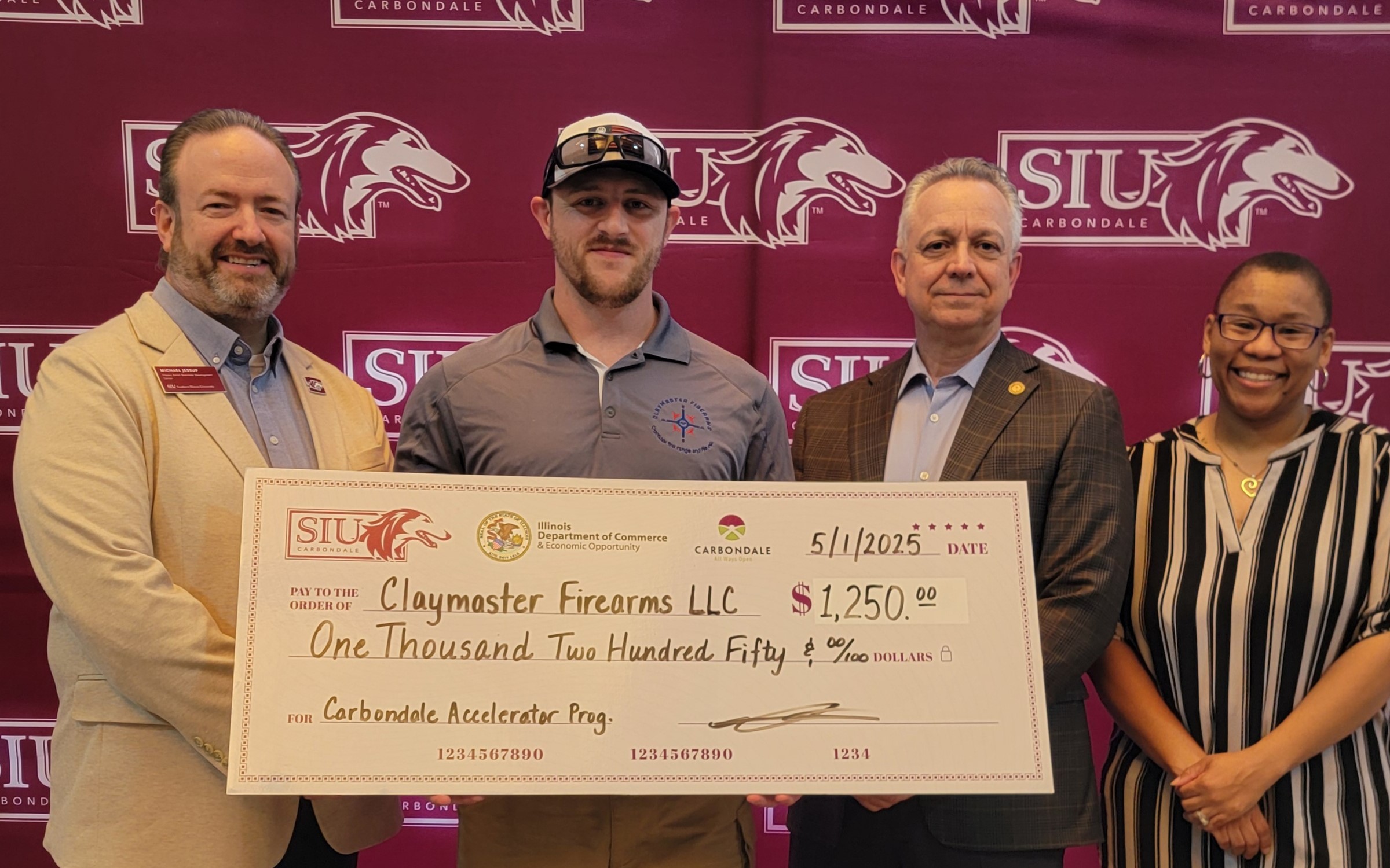 Matthew Huff, owner of Claymaster Firearms, holds a ceremonial check at the Carbondale Accelerator Program graduation ceremony. SBDC advisors and Steve Mitchell from the City of Carbondale stand on either side of him. 