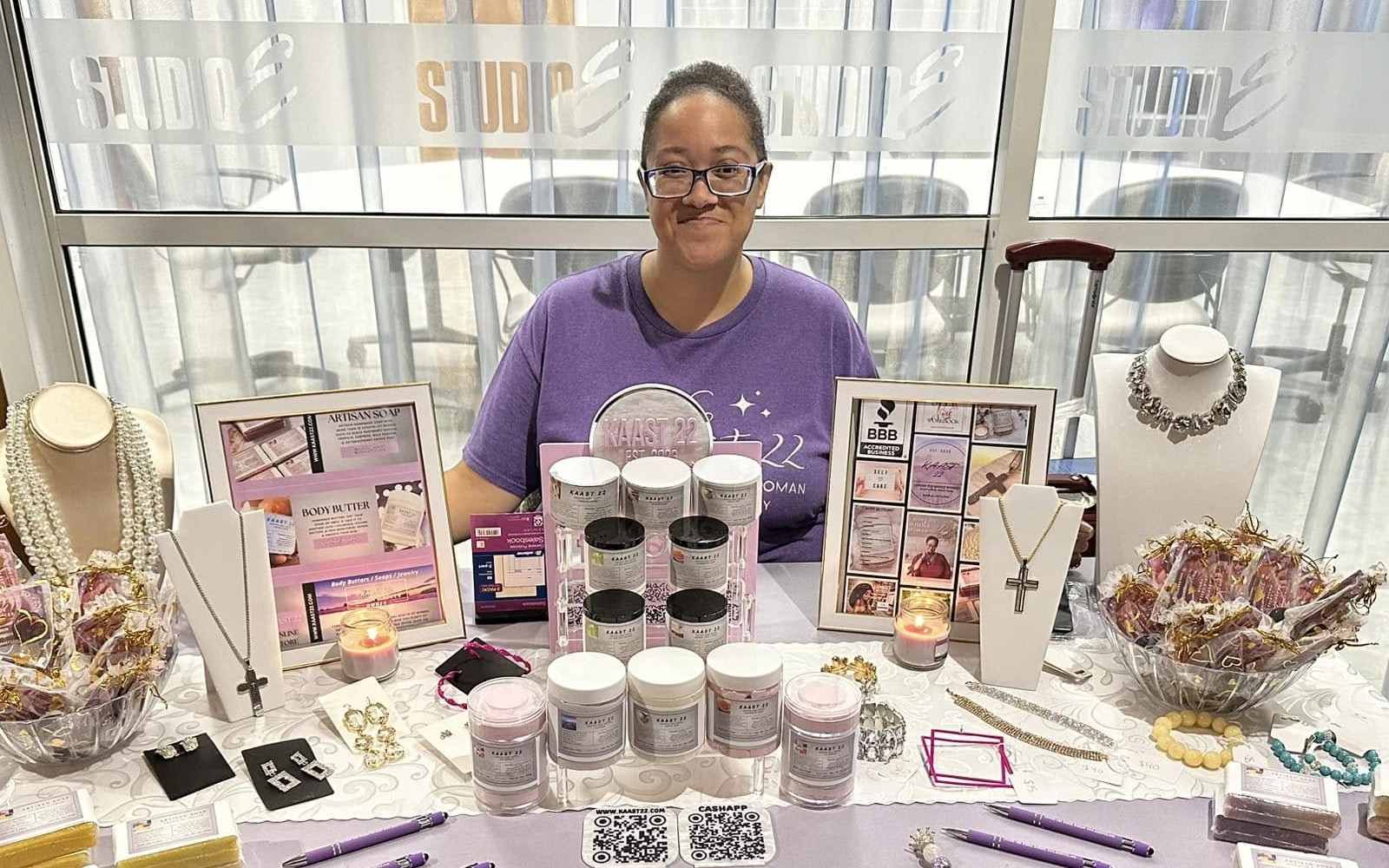 Carla Casler sits behind her display table at a vendor fair. Her table is decorated with body butters, soaps, and jewelry. 
