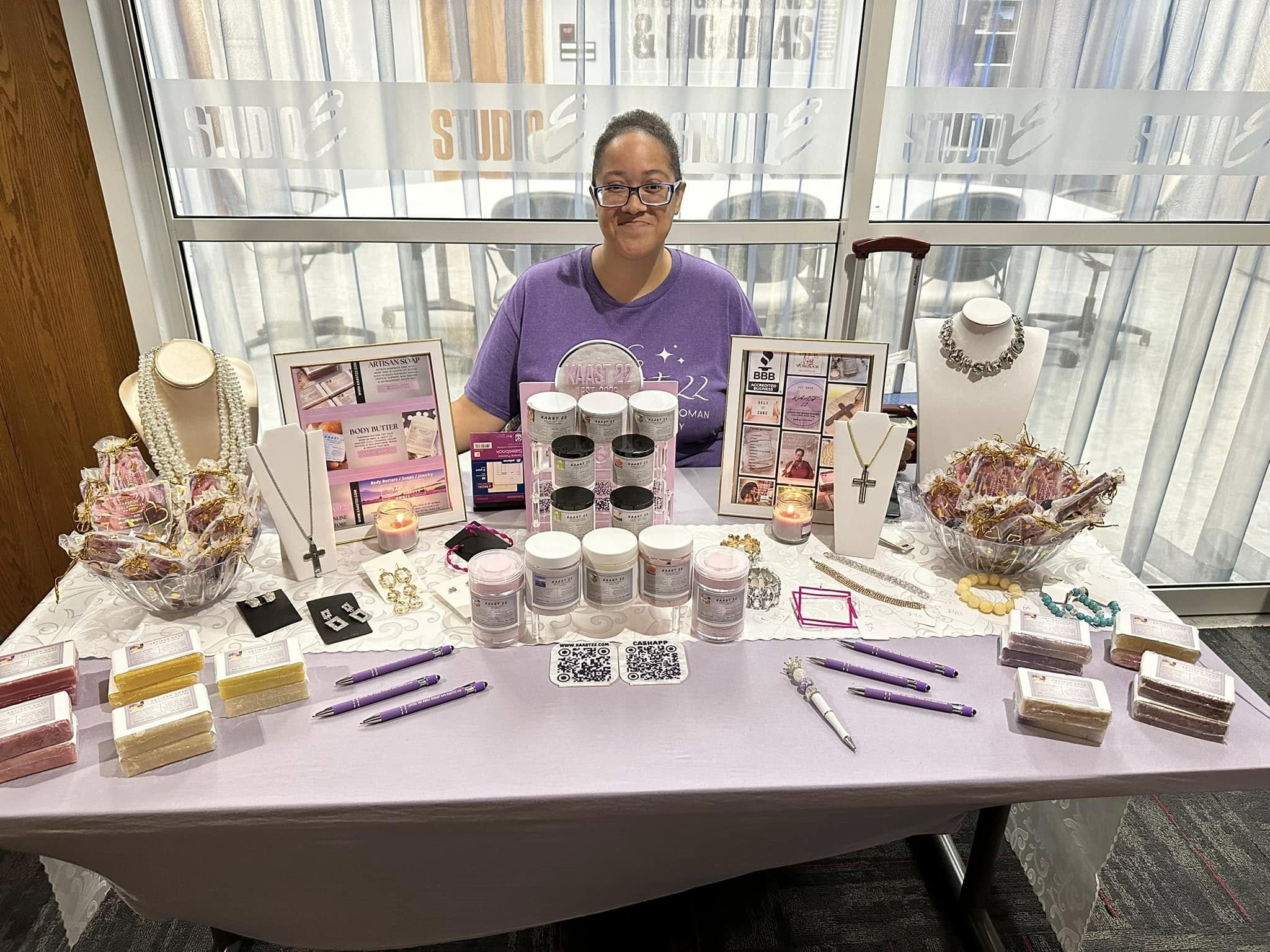 Carla Casler sits behind her display table at a vendor fair. The table is decorated with body butters and lotions. 